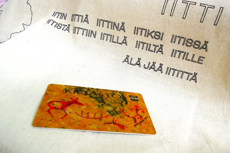 Kuva kuuluu näyttelyaineistoon. Siinä on viittaukset Iitin kunnallisten kansakoulujen opetukseen ja Iitin alueen esihistoriallisiin kalliomaalauksiin 2000-luvun alkuvuosina käytössä olleessa Iitin kirjastokortissa.